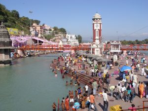 Image of the River Ganges in Haridwar, India. To the right there are people gathering on the banks and bathing in the river. A bridge and city buildings appear in the background.