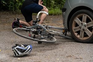 Image of a car striking a bike on the road. The biker is in the background of the image on the ground.