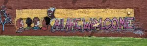 Mural on a brick wall of a teacher and several children of different races. "All are welcome" written across mural.