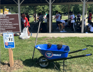 A wheelbarrow, gardening gloves, and rake sit in front of dozens of student volunteers at a park.