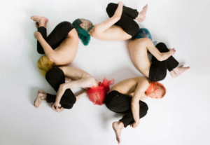 Five men lay on their sides in a circle, each blindfolded by a colorful ceremonial scarf and holding his knees to his chest.