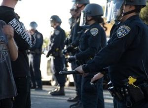 Law Enforcement Officers wearing full uniforms and holding batons face demonstrators.