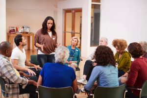 A woman stands as she leads a conversation among ten sitting community members.