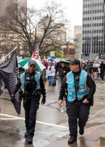 Two police officers in blue bests working with demonstrators at a protest in a downtown, urban area.