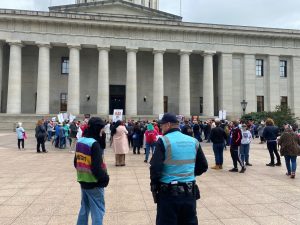 A law enforcement officer wearing a blue vest speaks with a demonstrator during a protest in a public space.