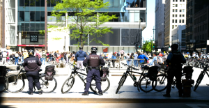 Law enforcement officers on bicycles near demonstrators in a downtown, urban area.