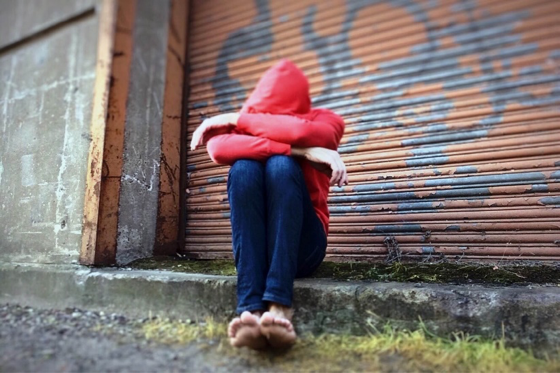 person in red hoodie with head down, arms crossed, and seated in front of a graffit'di door