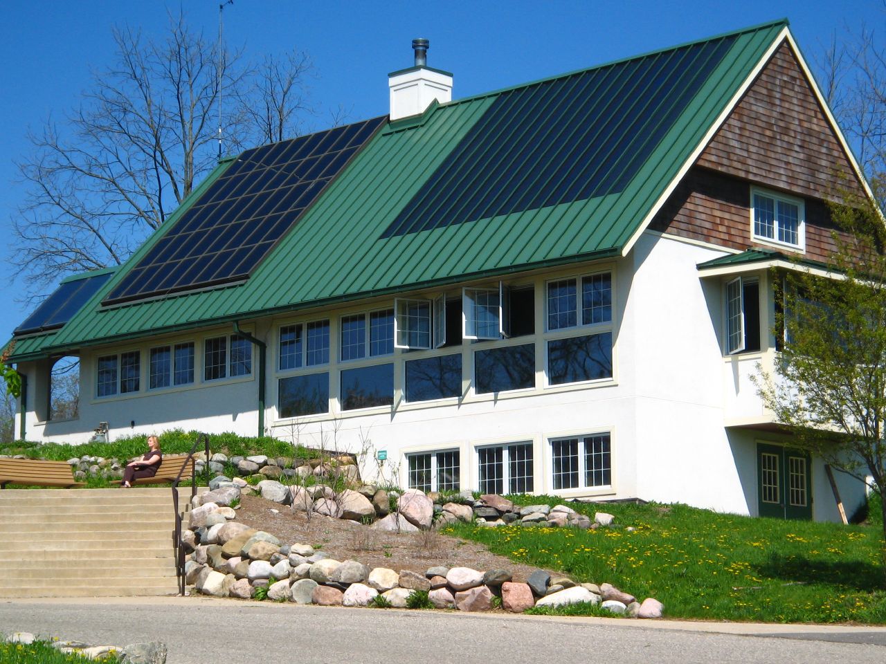 house with roof showcasing a standard solar panel with many separate cells on top of the roof and a one panel that mimics the long, straight roof tiles.